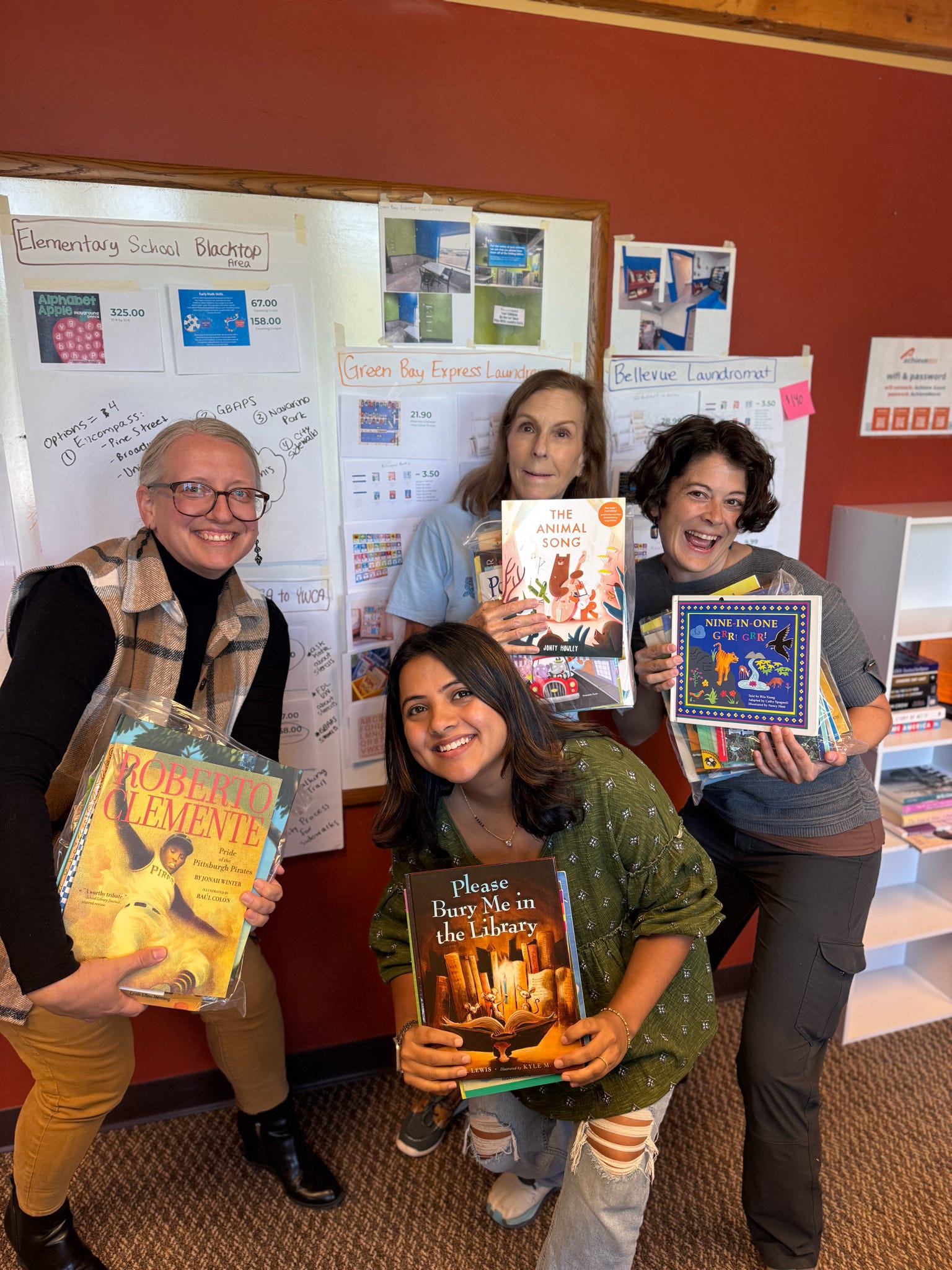 Four individuals in a library setting holding various children's books, smiling and posing for the camera. A bulletin board in the background displays information about local services.