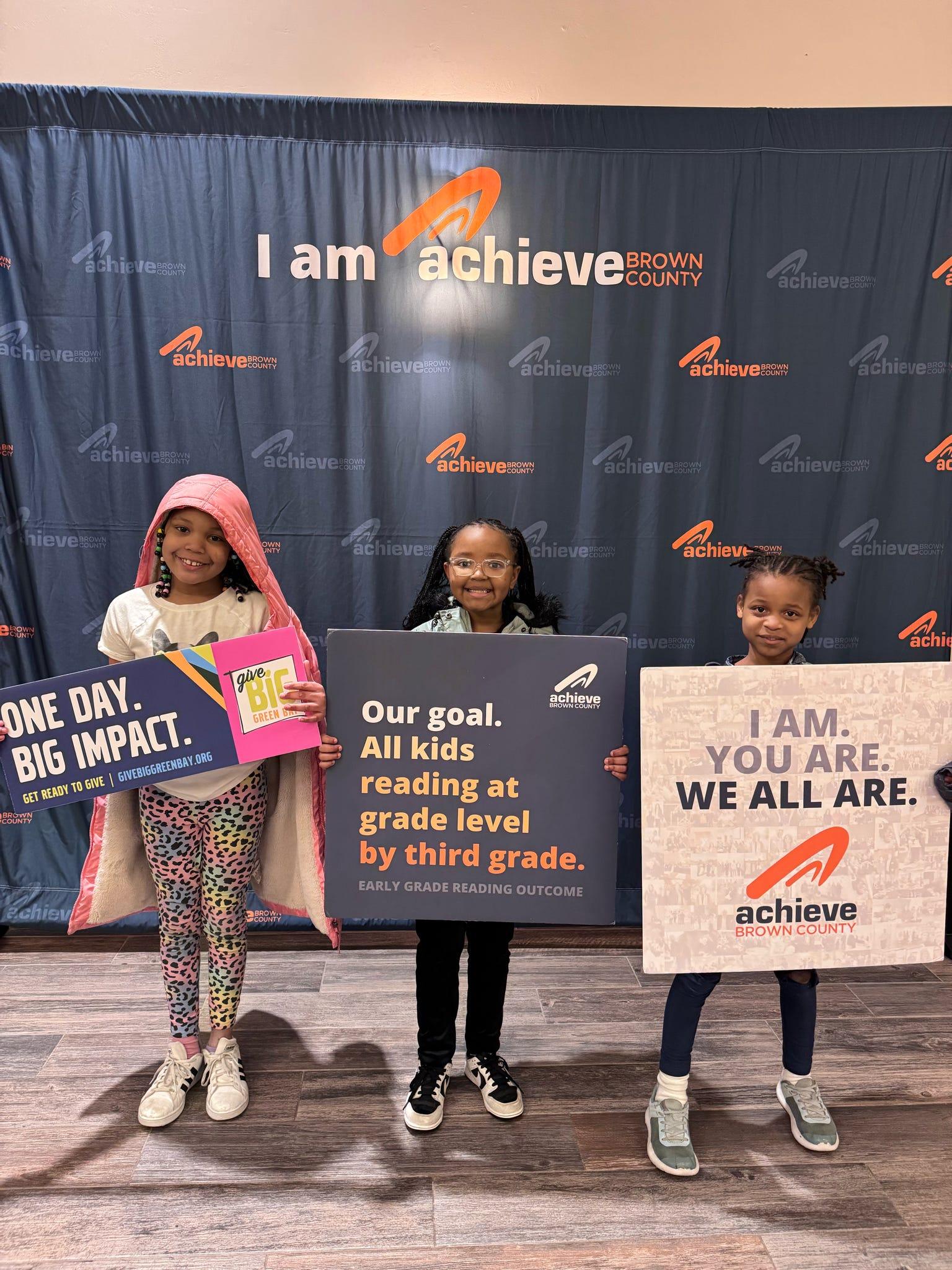 Three children standing in front of a banner that says 'I am achieve Brown County.' They are holding signs promoting early grade reading outcomes. One sign states 'Our goal. All kids reading at grade level by third grade.' The other reads 'One Day. Big Impact.'