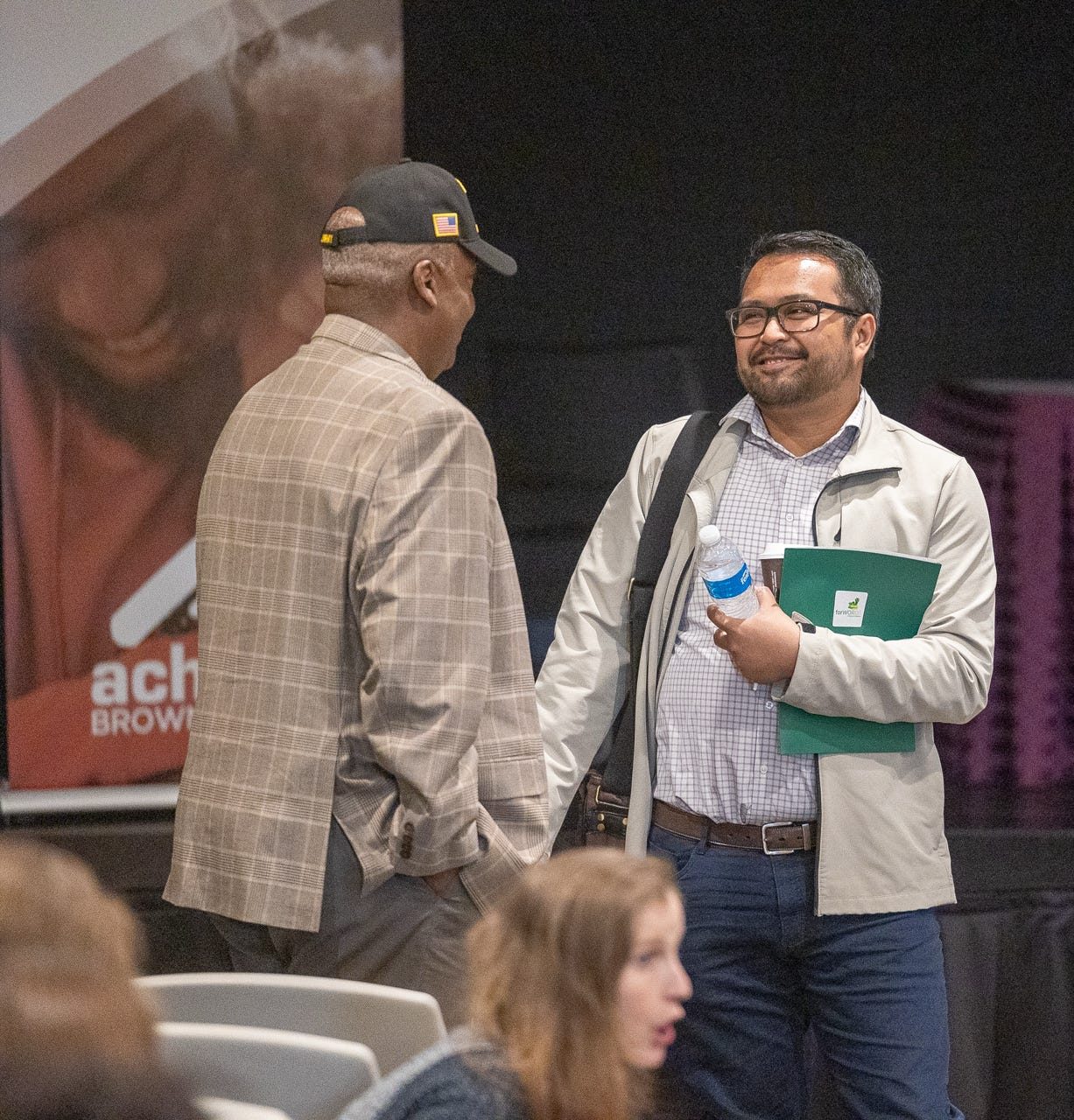 Two men engaged in conversation at an event, one holding a water bottle and documents, with a group of people seated in the background.